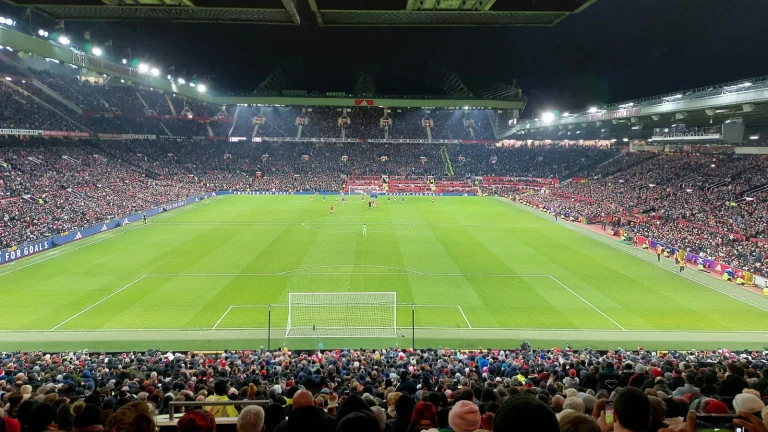 Volgepakte tribunes in Old Trafford tijdens een avondwedstrijd, gezien vanaf de Stretford End met supporters op de voorgrond